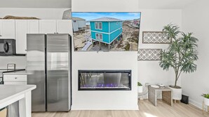 Eine moderne Küche mit weißen Schränken, einem Edelstahlkühlschrank, einem eingebauten Kamin und einem an der Wand montierten Fernseher, der ein Haus an der Küste zeigt.