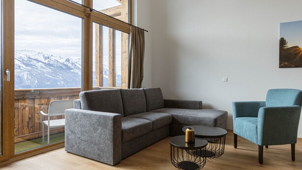 Un salon moderne avec un canapé d’angle gris, une table ronde noire ornée d’un vase et un fauteuil bleu. De grandes fenêtres offrent une vue sur des montagnes enneigées.