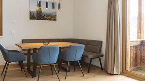 Un coin repas avec une table en bois, des chaises bleues et un banc. Il y a un tableau au mur et une fenêtre avec des rideaux.