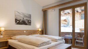 Une chambre avec une tête de lit en bois, deux lampes de chevet, un grand lit avec du linge de lit blanc, un banc en bois, une porte-fenêtre coulissante donnant sur un espace extérieur et un tableau représentant un paysage de montagne accroché au mur.