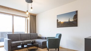 Un salon moderne avec un canapé d’angle gris, un fauteuil bleu, deux petites tables rondes noires et une grande fenêtre avec des rideaux.