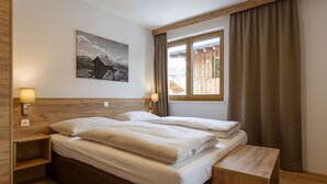 A double bed with a wooden headboard, two bedside tables with lamps, a wooden bench, a large window with curtains, and a black and white landscape painting on the wall.