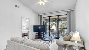 Una sala de estar con televisión, dos sofás, una mesa de centro de vidrio y un balcón con mobiliario exterior.