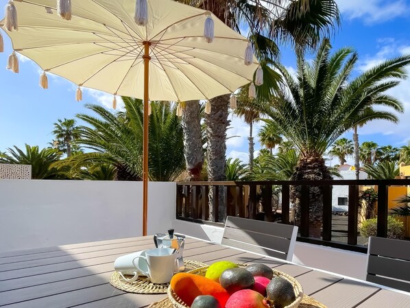 A patio with a table set for two, a coffee maker, and a basket of fruit.