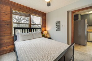 A bedroom with a wooden headboard, a large window, a bed with white bedding, a nightstand with a lamp, and a kitchen visible through an open door.