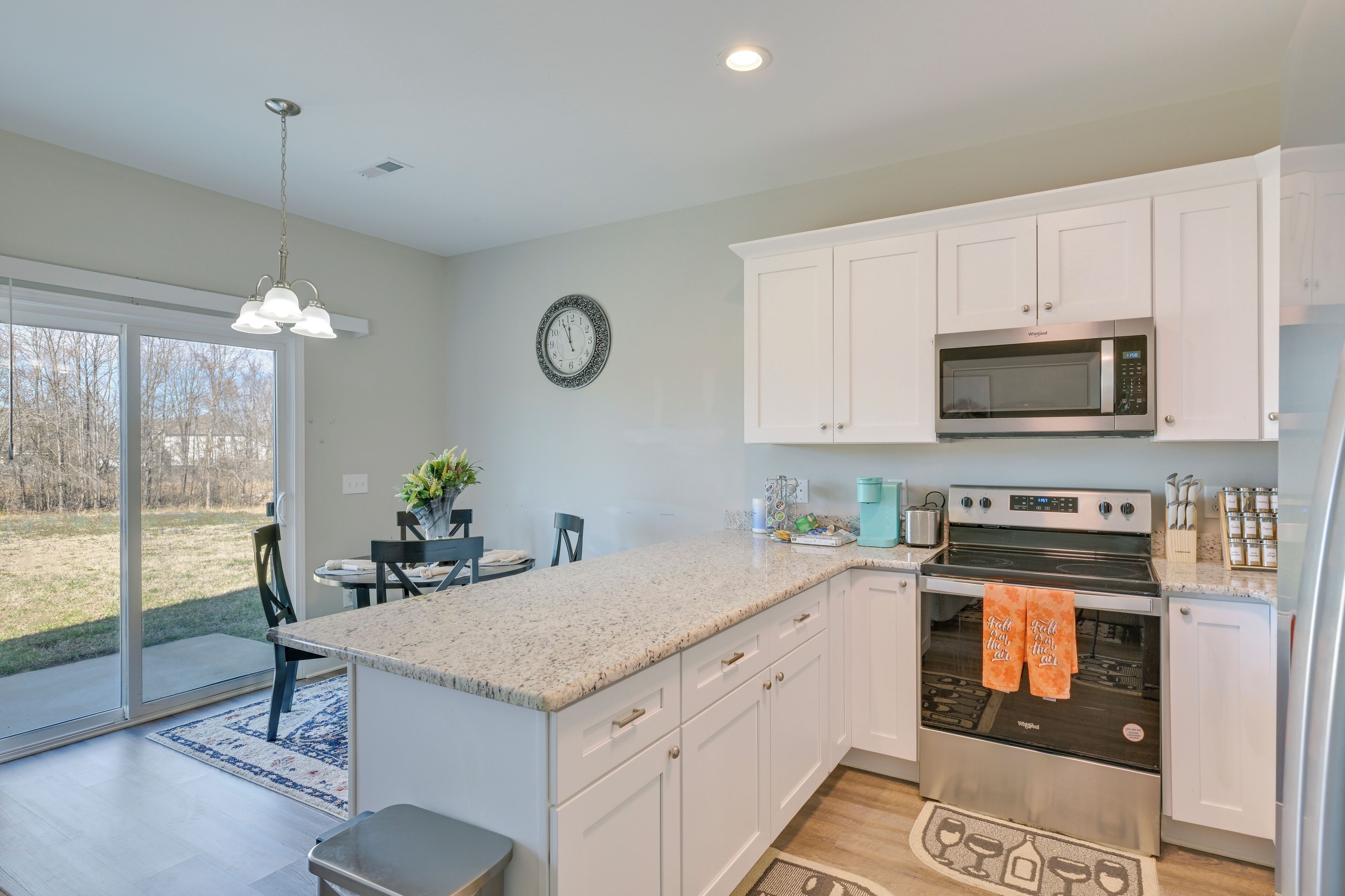 A modern kitchen with white cabinets, a stainless steel oven, and a microwave. There is a dining area with a table and chairs, and a large window providing a view of the outdoors.