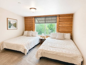 A room with two beds, a window with wooden shutters, a framed picture on the wall, and a small table with a lamp.