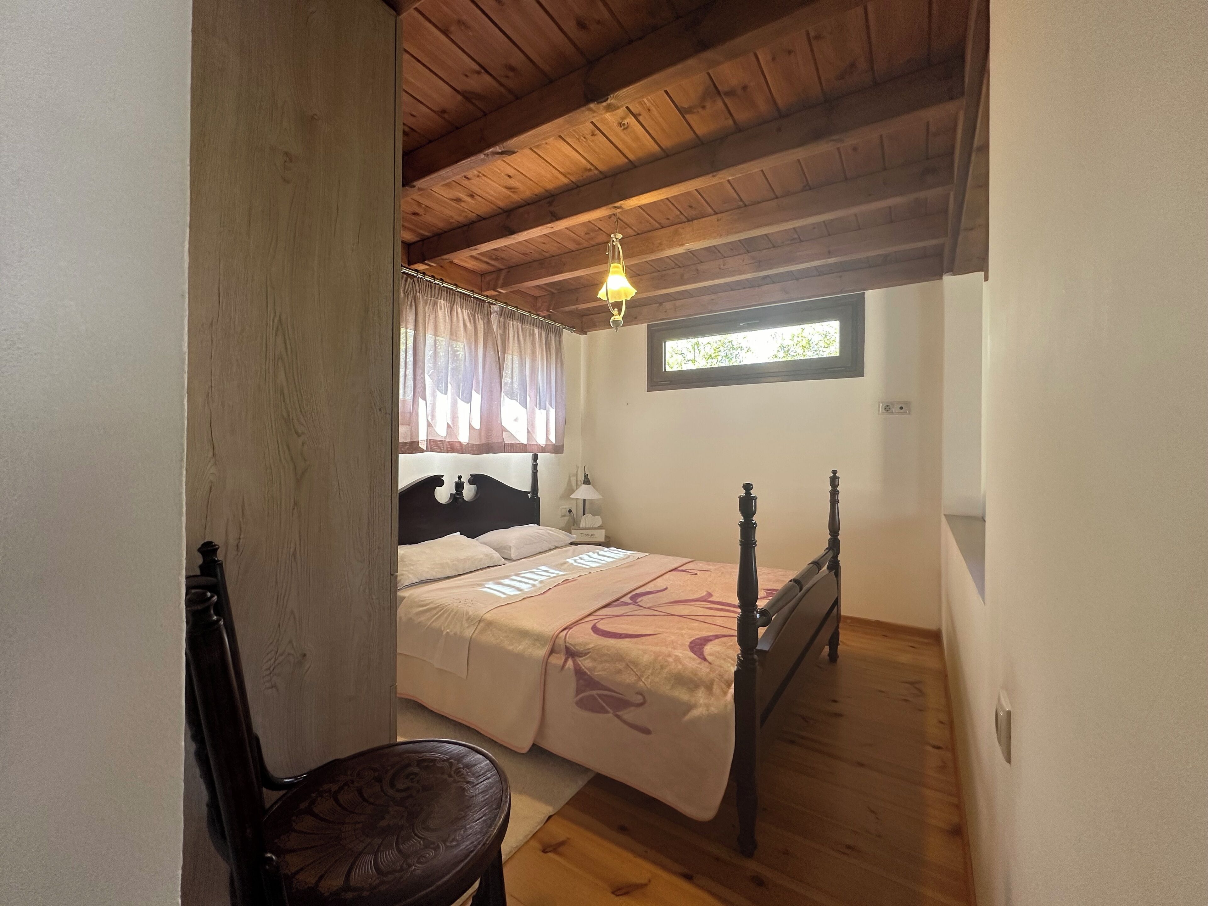 Un dormitorio con techo de madera, una cama con cabecera, una mesita de noche, una silla y una ventana con cortinas.