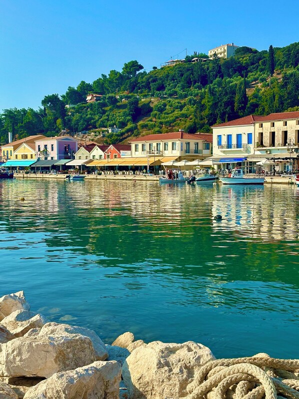 A coastal town with buildings along the waterfront, boats docked, and a hill with a house in the background.