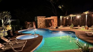 Un área de piscina nocturna con muro de piedra, sillones de descanso y una pequeña cascada.