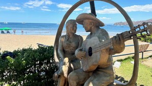 Escena de playa con una estatua de un hombre tocando la guitarra y una mujer sentada a su lado, rodeados de follaje.