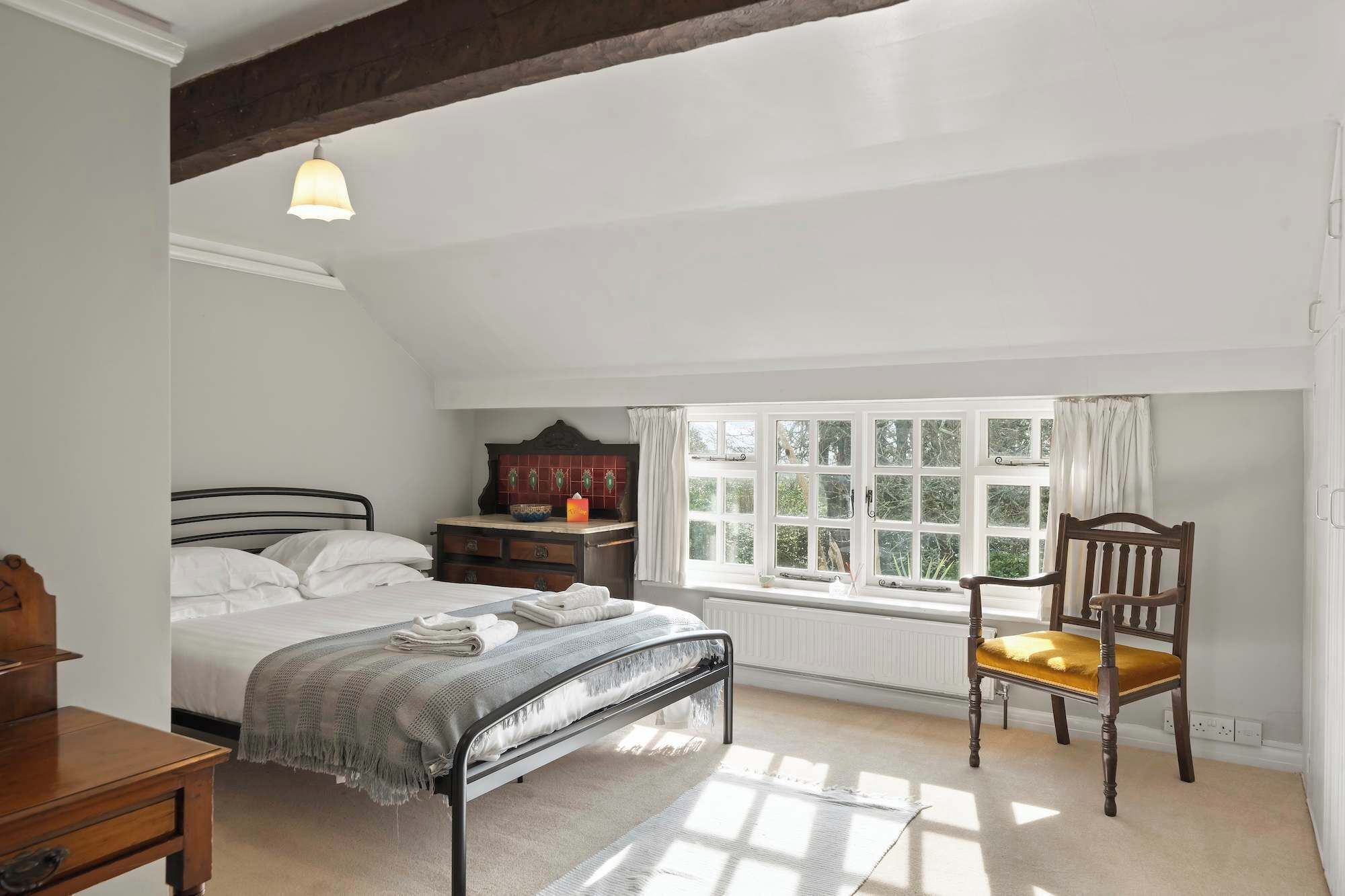 Une chambre à coucher avec un lit, une chaise en bois, une commode, une fenêtre avec des rideaux et un radiateur.