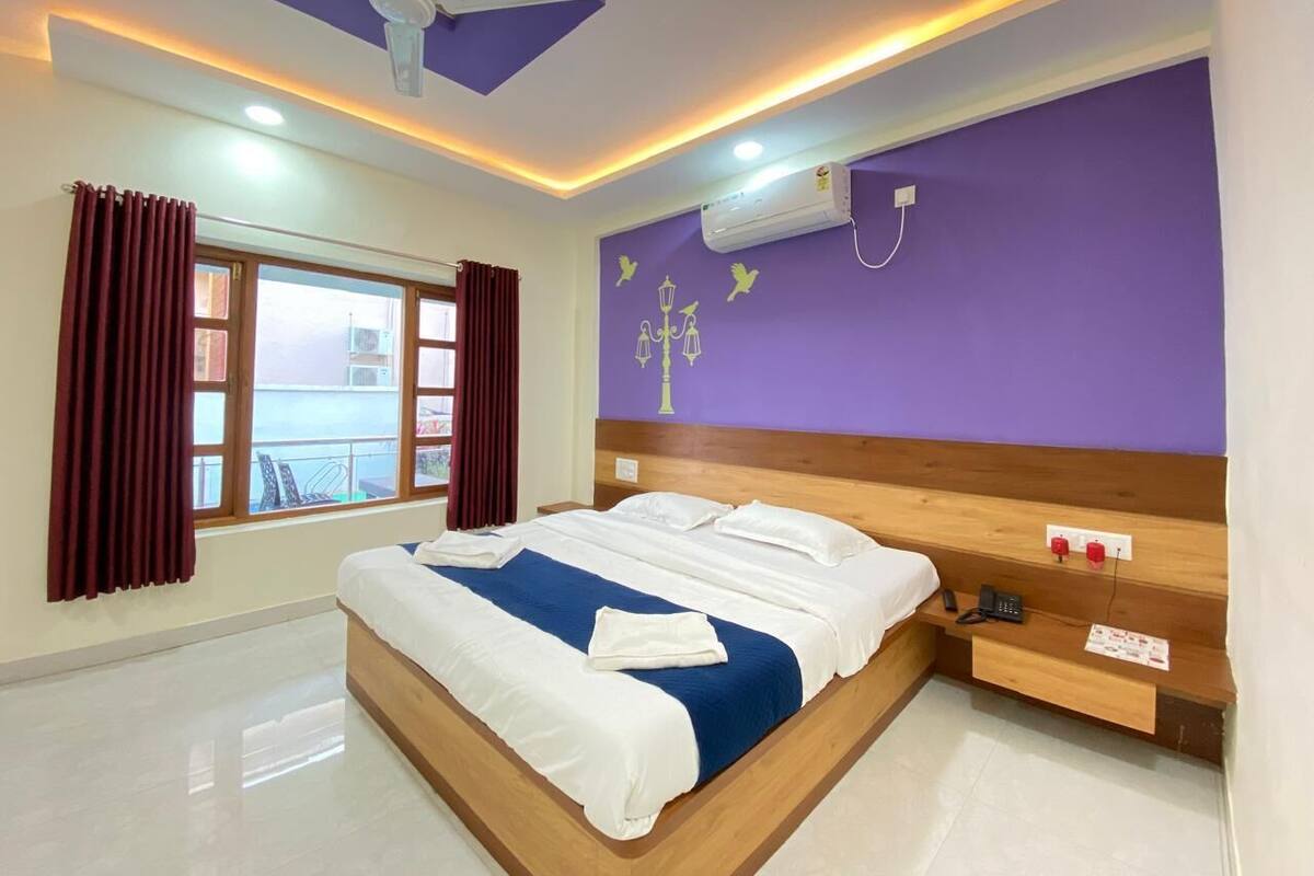 Ein modernes Hotelzimmer mit einem großen Bett, einem Holz-Kopfteil, einem Nachttisch, einem Fenster mit Vorhängen und einem Deckenventilator.