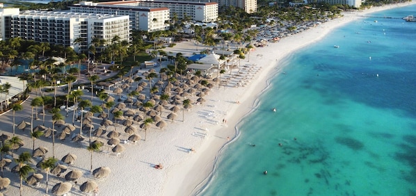 A beach resort with multiple hotel buildings, a sandy beach lined with palm trees and umbrellas, and clear turquoise waters.