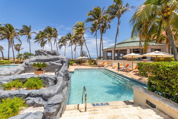 A pool area with a rock formation, palm trees, lounge chairs, and a building with a green roof.