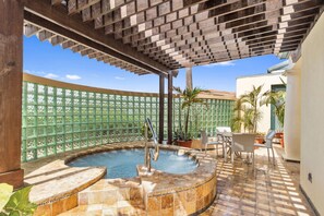 Un jacuzzi circulaire, bordé de carreaux, entouré d’une structure en forme de pergola. Des chaises et des plantes en pot sont disposées autour du jacuzzi.