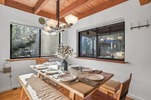 A dining area with a wooden table set for four, large windows, and a ceiling with exposed beams.