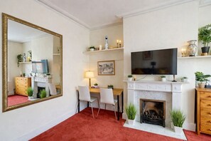 Quarto de hotel com um espelho grande, lareira, televisão, escrivaninha com cadeiras e um tapete vermelho.