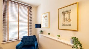 Um quarto com uma cadeira azul, uma janela com persianas, um abajur e duas molduras com quadros na parede.