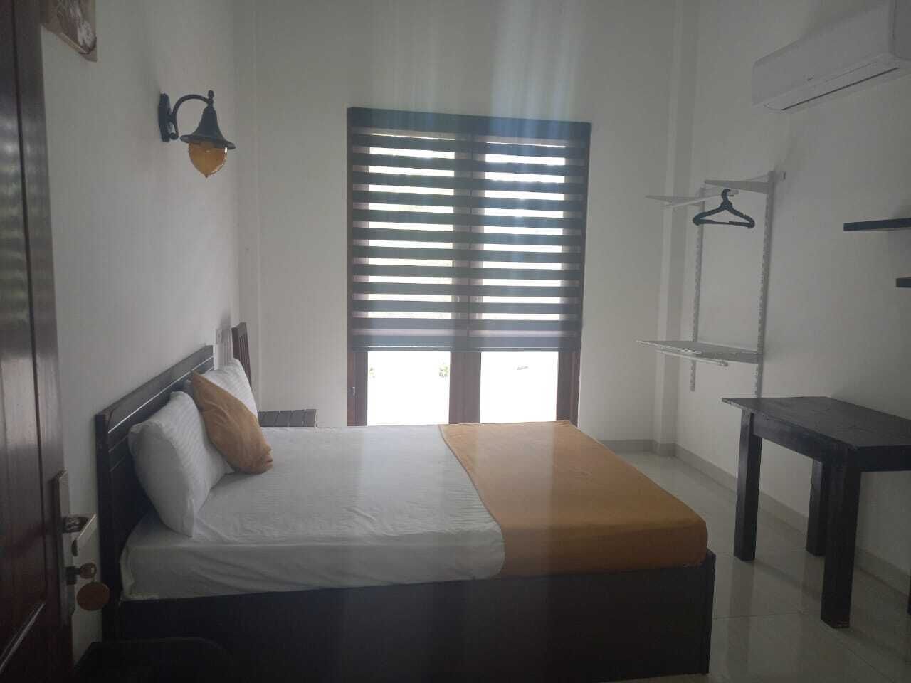 A bedroom with a bed, a bedside table, a hanging rack, and a window with blinds.