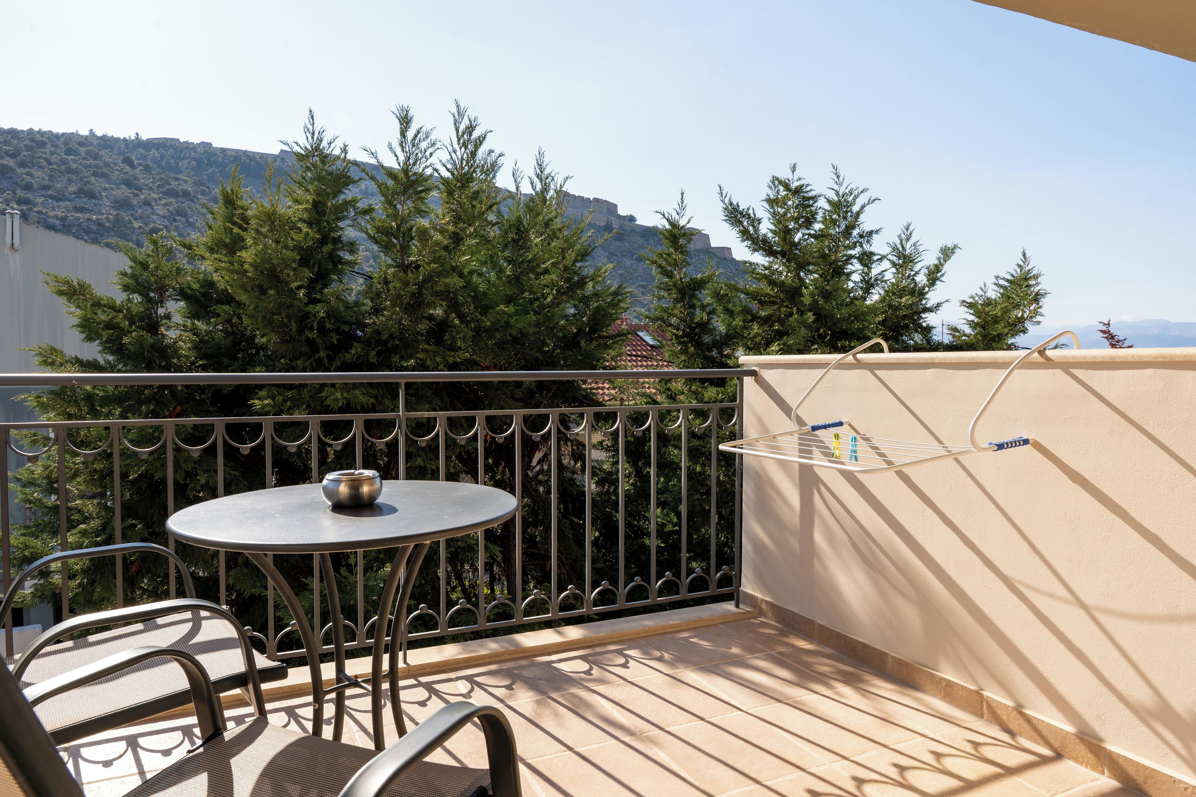 A balcony with a round table and chairs, a drying rack, and a view of a hill with trees and a building in the background.