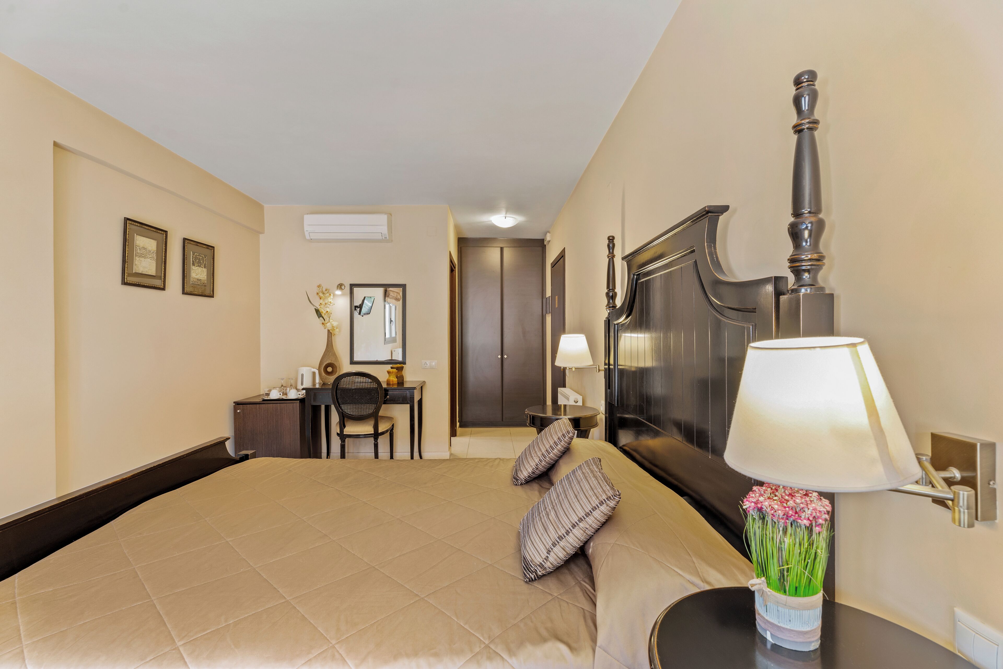 Habitación de hotel con una cama grande, un escritorio con silla, una lámpara y flores, y un armario.