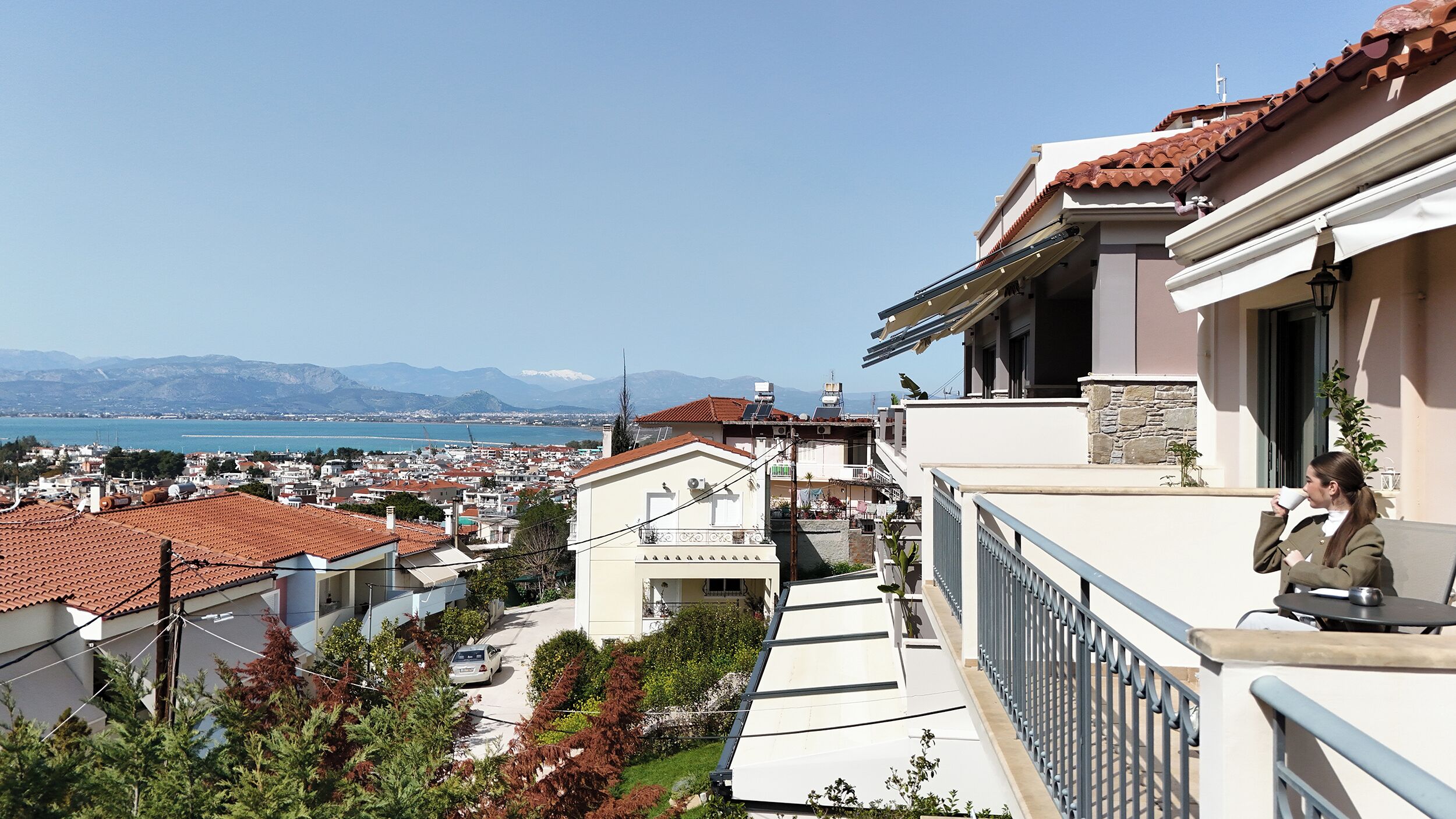 A residential area with a view of the sea and mountains. There are houses with red-tiled roofs and balconies.