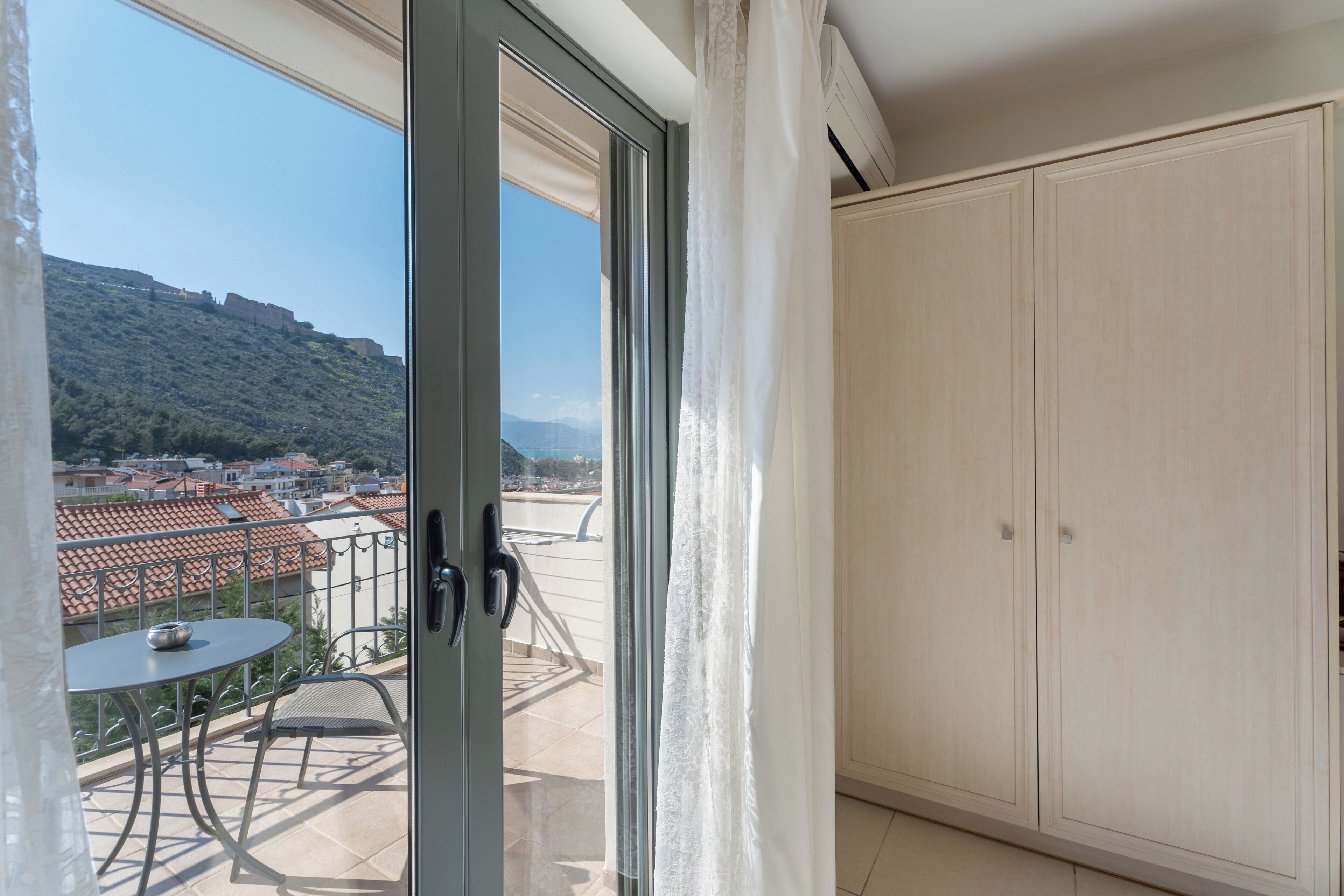 A balcony with a table and chairs, a sliding glass door, and a view of a town with buildings and a hill.