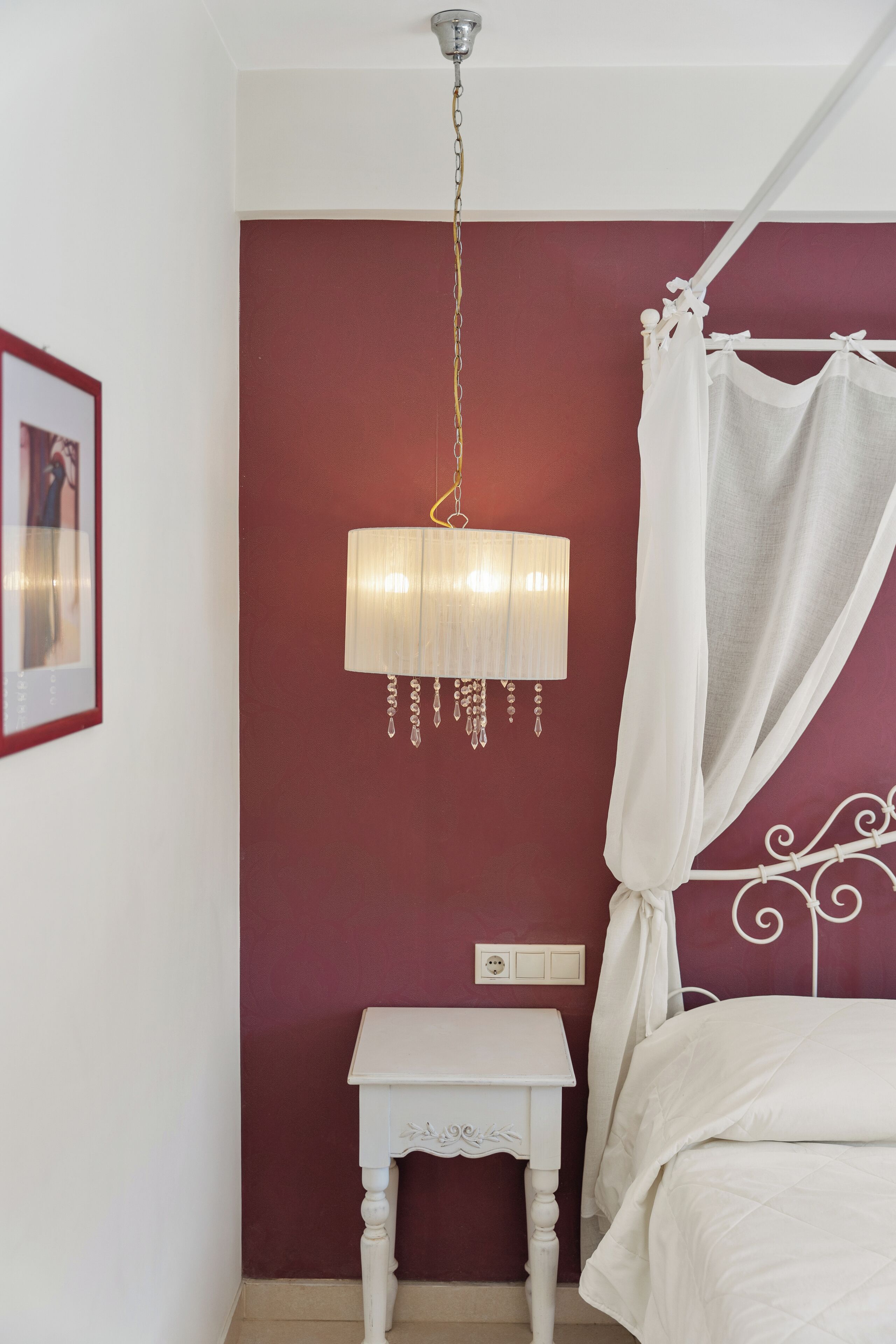 Une chambre avec un mur rouge, un lit blanc à baldaquin, une table de chevet et une lampe suspendue.