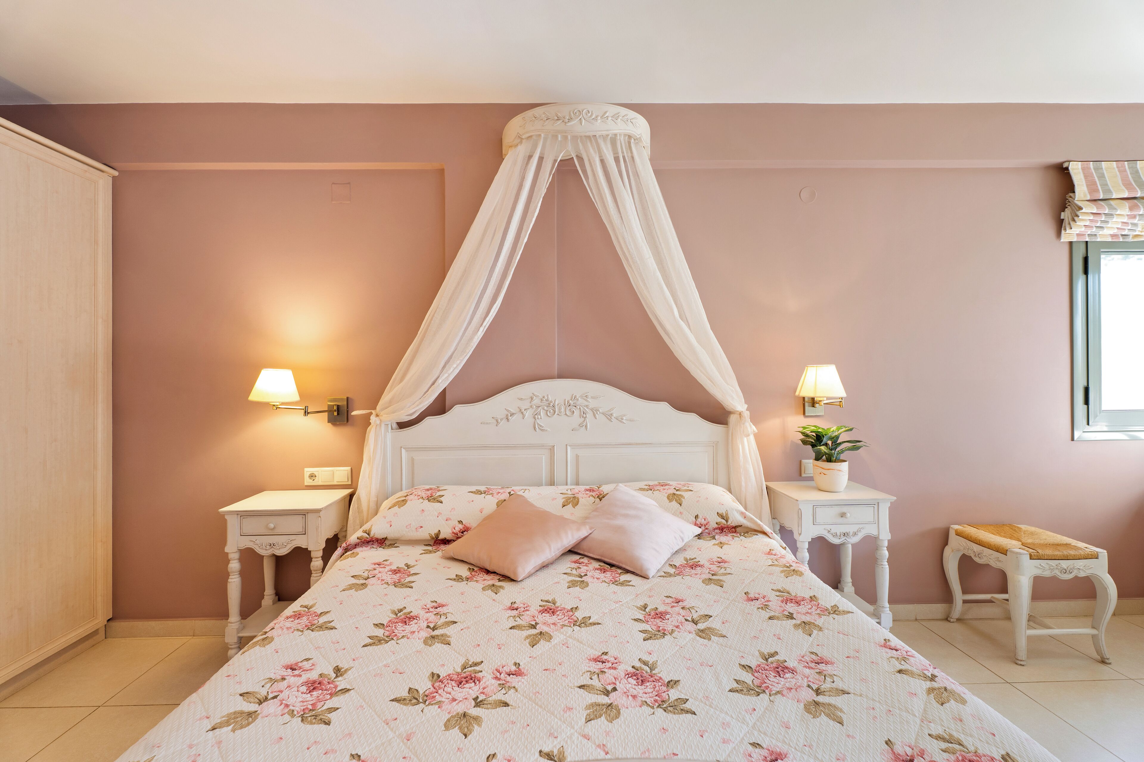 A bedroom with a canopy bed, bedside tables with lamps, a small bench, and a potted plant.