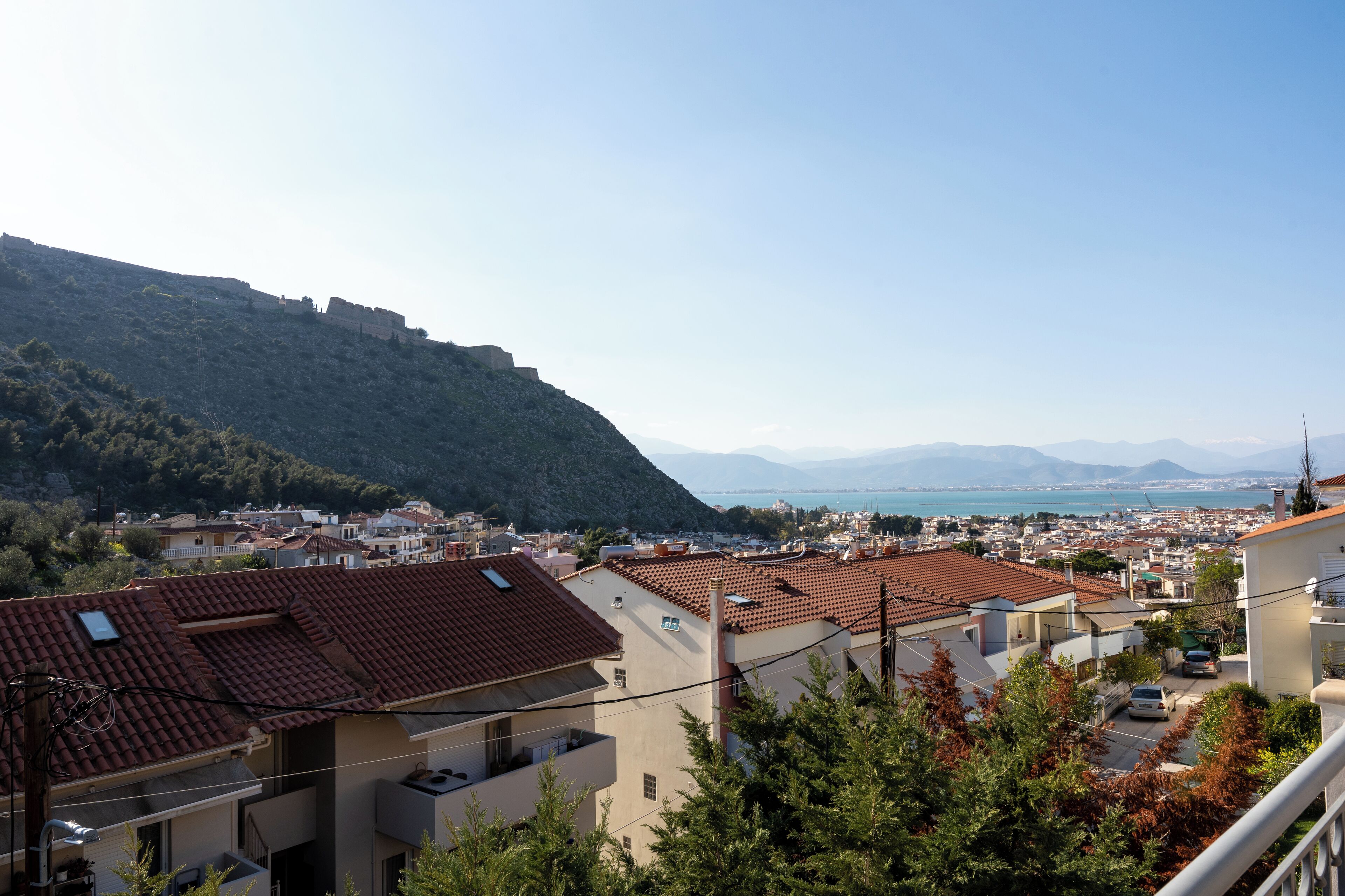 A coastal town with terracotta-roofed buildings, a view of the sea, and a mountainous backdrop.