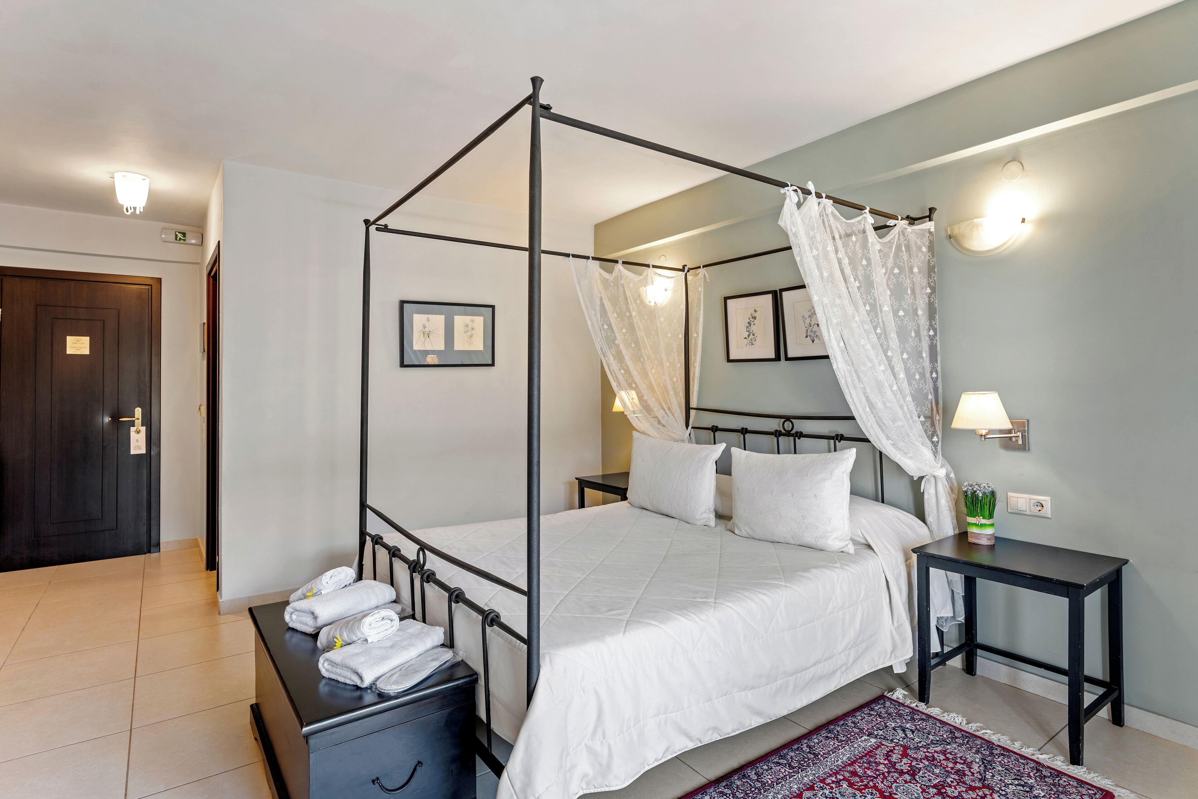 A four-poster bed with white bedding, a bedside table with a lamp, and a red patterned rug.