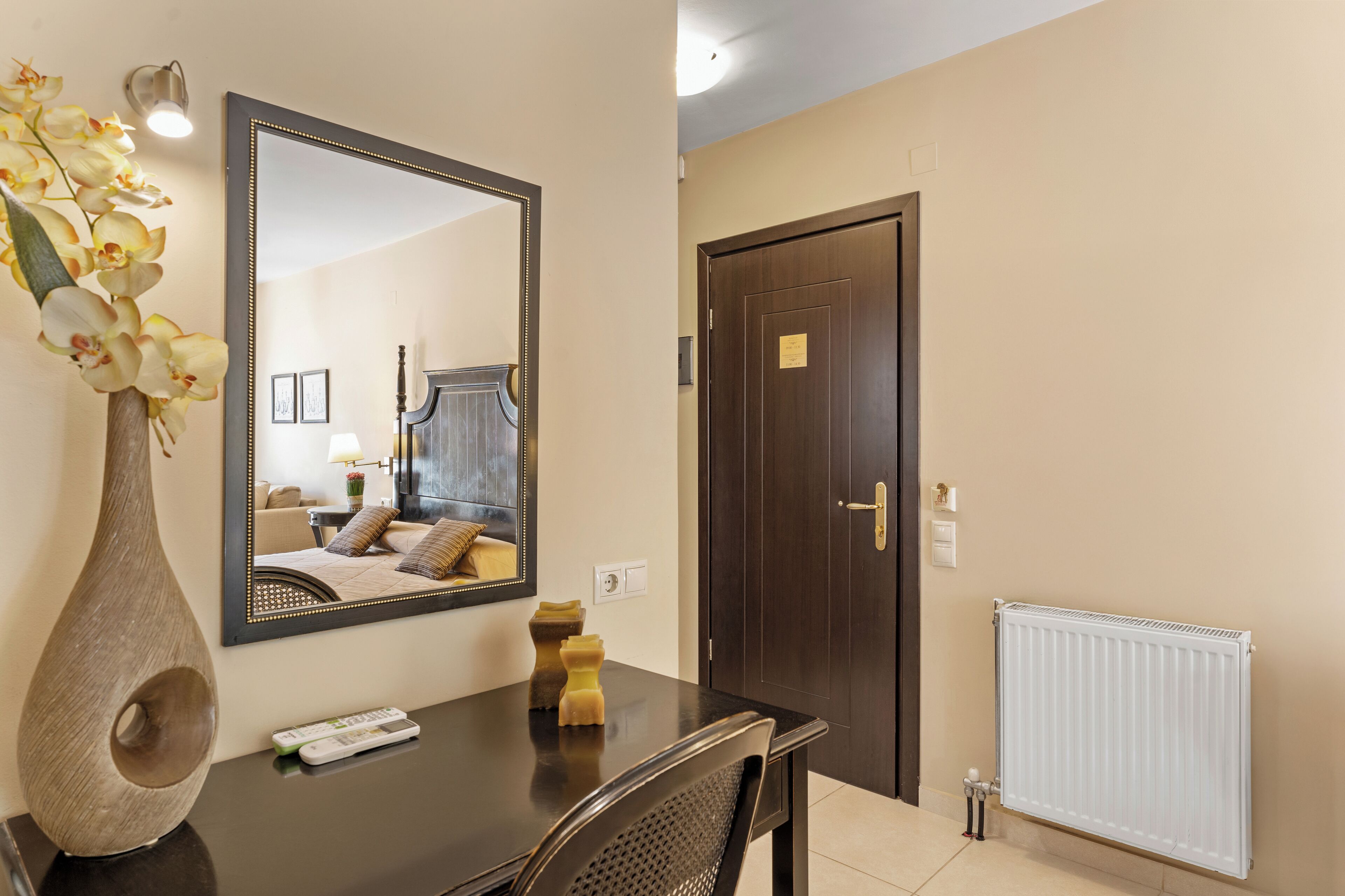Une chambre d’hôtel avec un bureau en bois foncé, un vase avec des fleurs, un miroir, une porte et un radiateur.