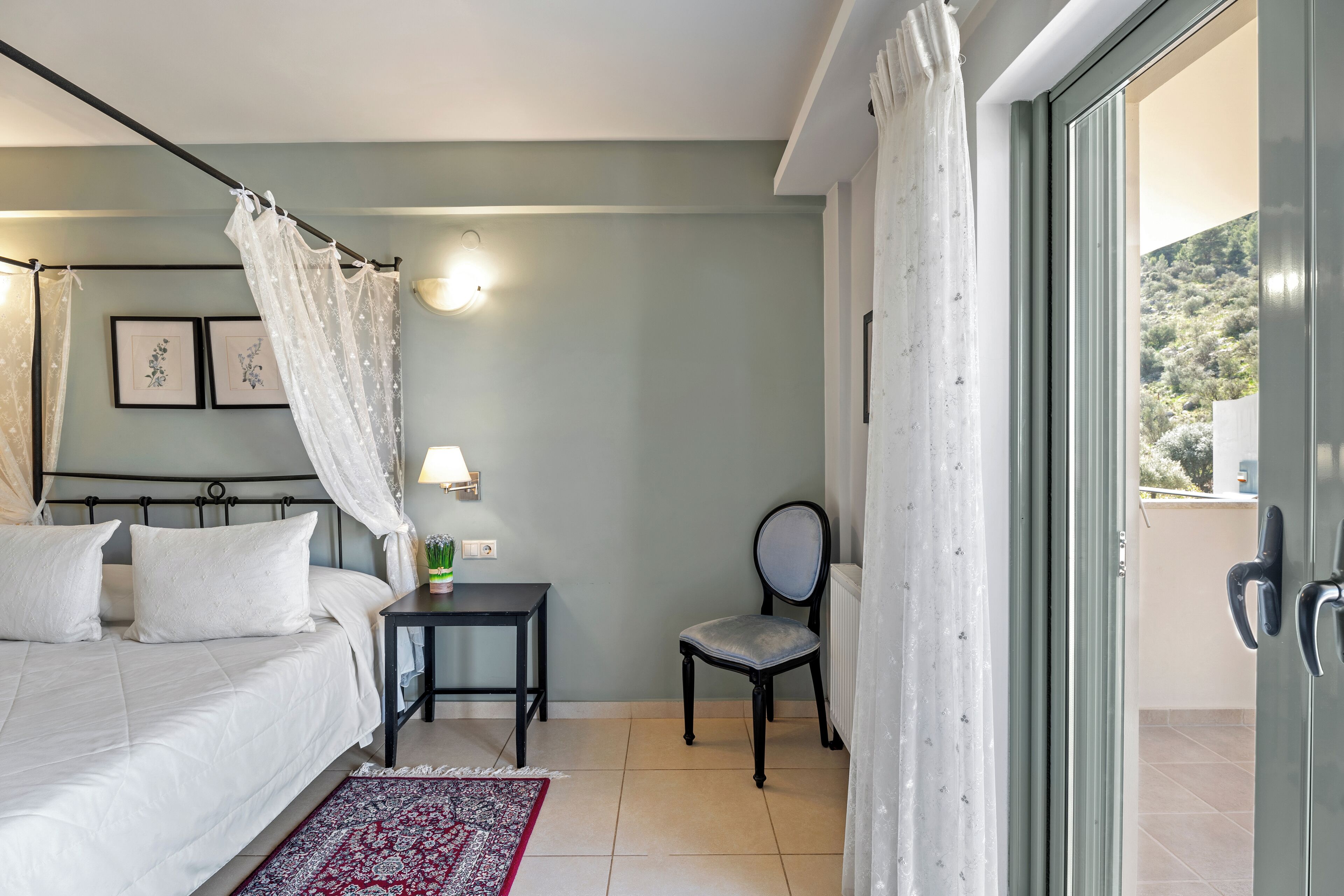 Une chambre à coucher avec un lit, une table de chevet, une chaise, une lampe et une vue sur l’extérieur, visible à travers une porte vitrée.