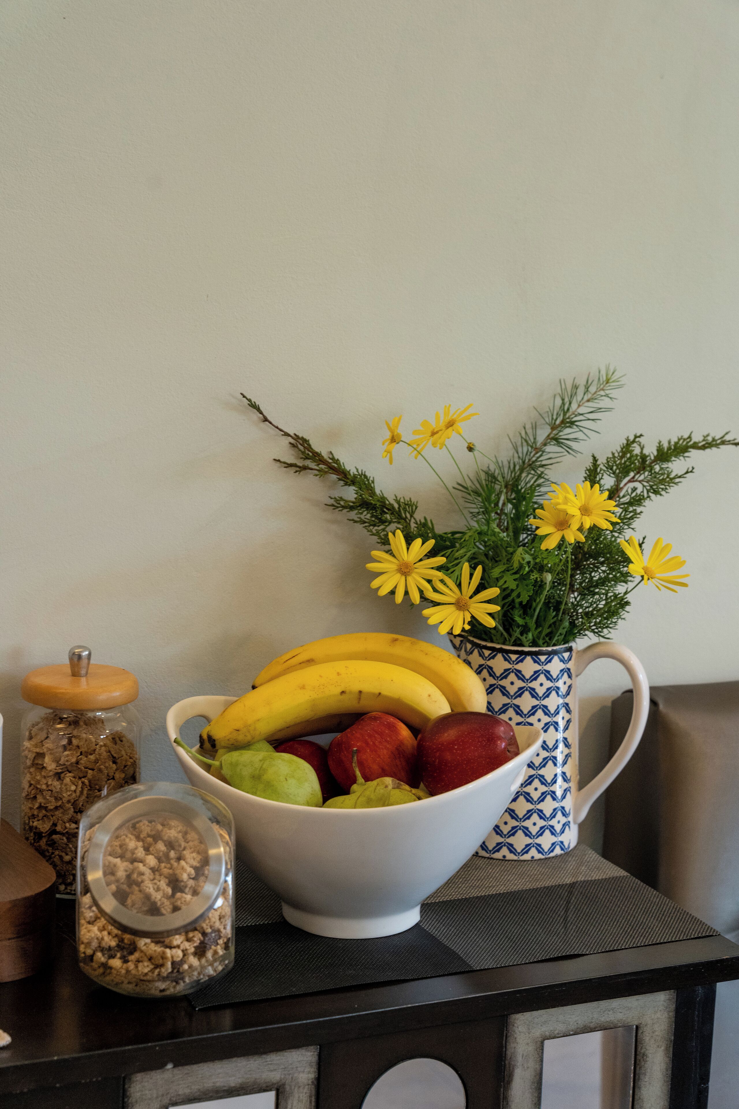 A table with a bowl of fruit, a jar of granola, and a vase with flowers.