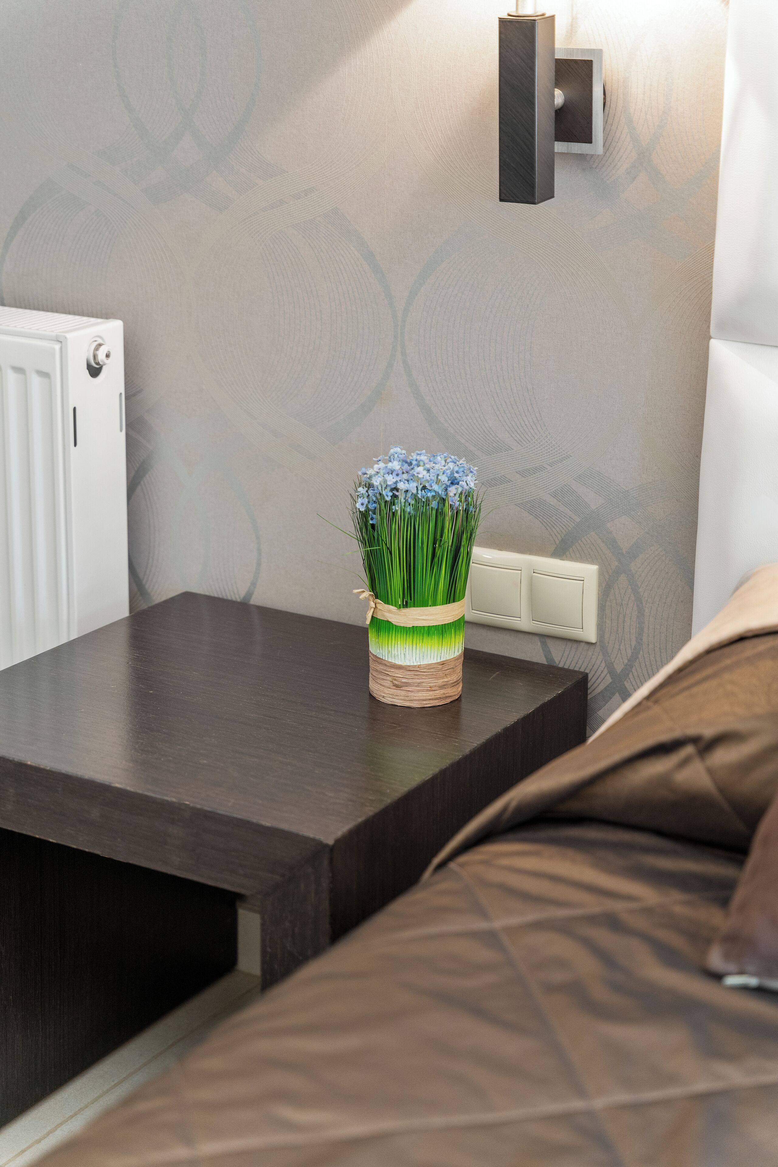 A neatly made bed with a brown cover, a bedside table with a vase of flowers, and a wall-mounted light fixture.
