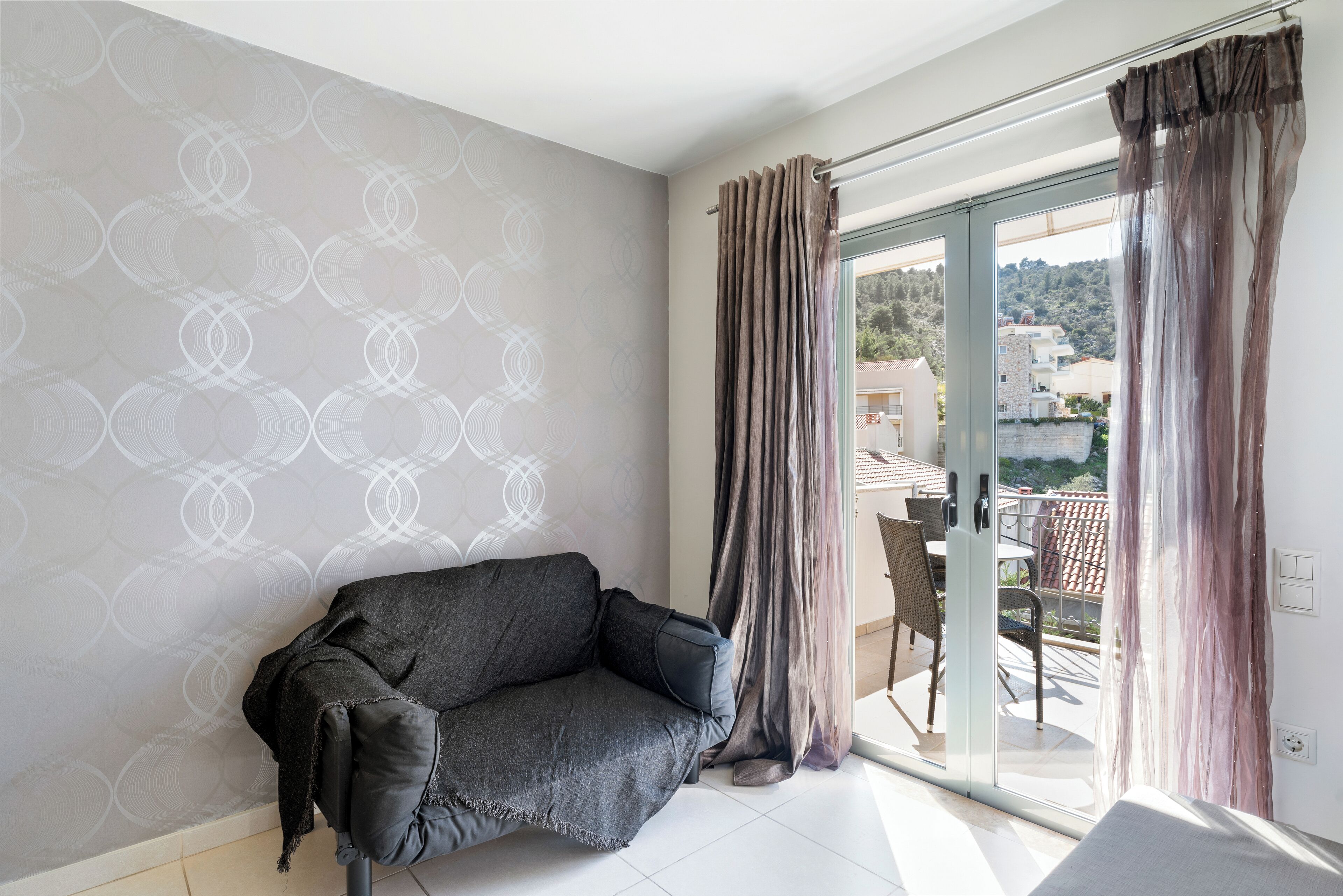 A modern living room with a grey sofa, a patterned wall, and a sliding glass door leading to an outdoor area with a table and chairs.