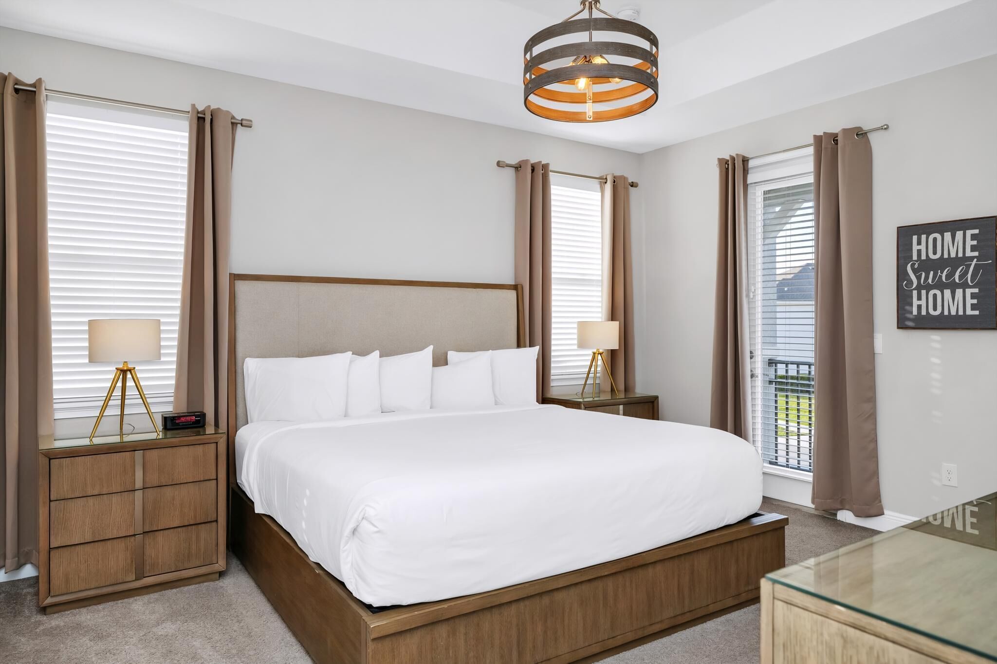 A bedroom with a large bed, two bedside tables with lamps, a modern chandelier, and a view of the outside.