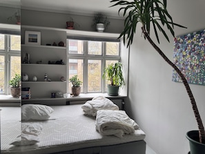 Quarto com cama, janela com vista, prateleira com objetos decorativos e planta em vaso.