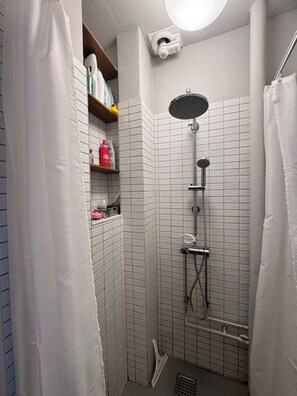 En liten, kaklad dusch med ett duschmunstycke som kan hållas i handen och en väggmonterad blandare.