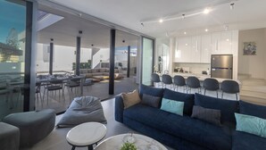 A modern living room with a blue sofa, a round coffee table, and a glass dining area. - Protaras Aqua Pearl Villa Aq18 (Paralimni)
