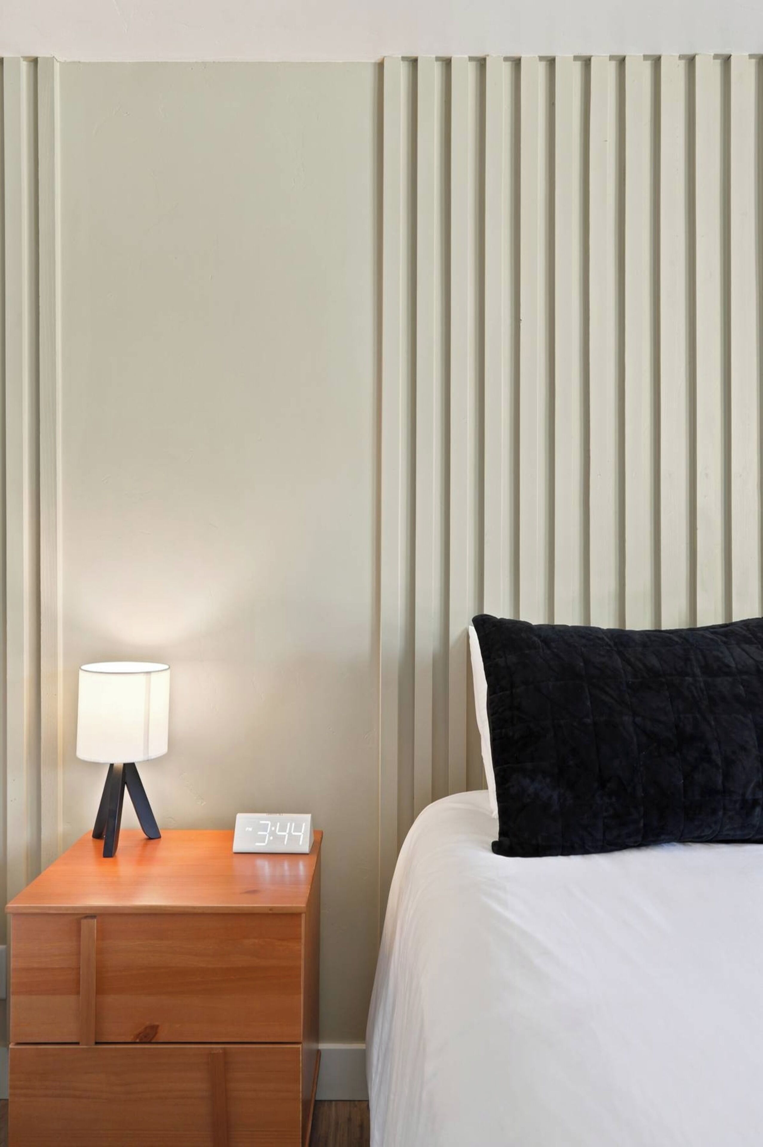 En pænt redt seng med en sort pude, et natbord med en lampe, og en væg med lodrette persienner.