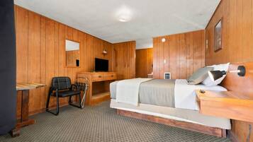 Una habitación con paneles de madera, una cama, un escritorio con una silla, un televisor y una pequeña mesa.