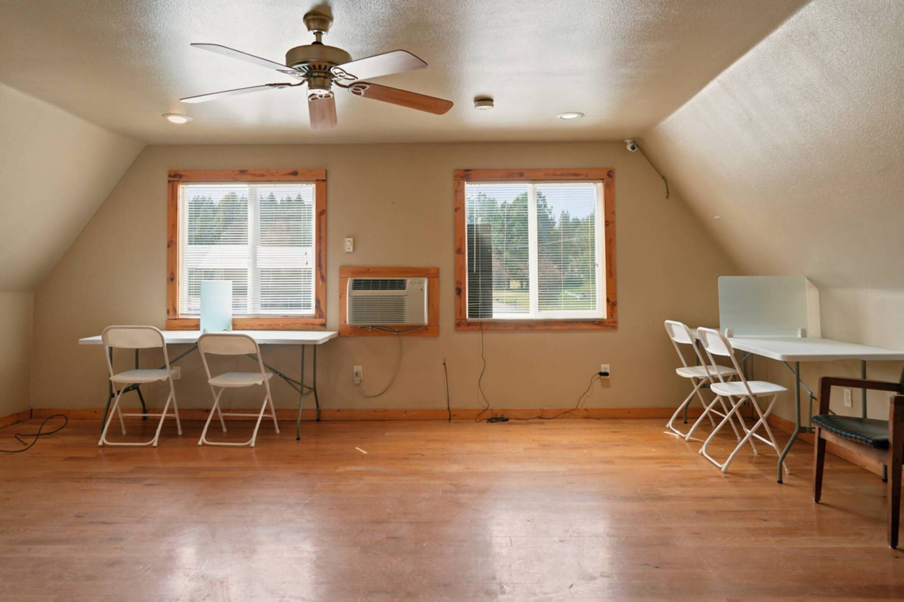 A room with a ceiling fan, two windows with blinds, a desk with a chair, and an air conditioning unit.