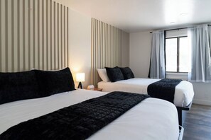 A hotel room with two beds, a nightstand with a lamp, a window with curtains, and a wall with vertical blinds. - Birch Glen Hotel (Cascade)