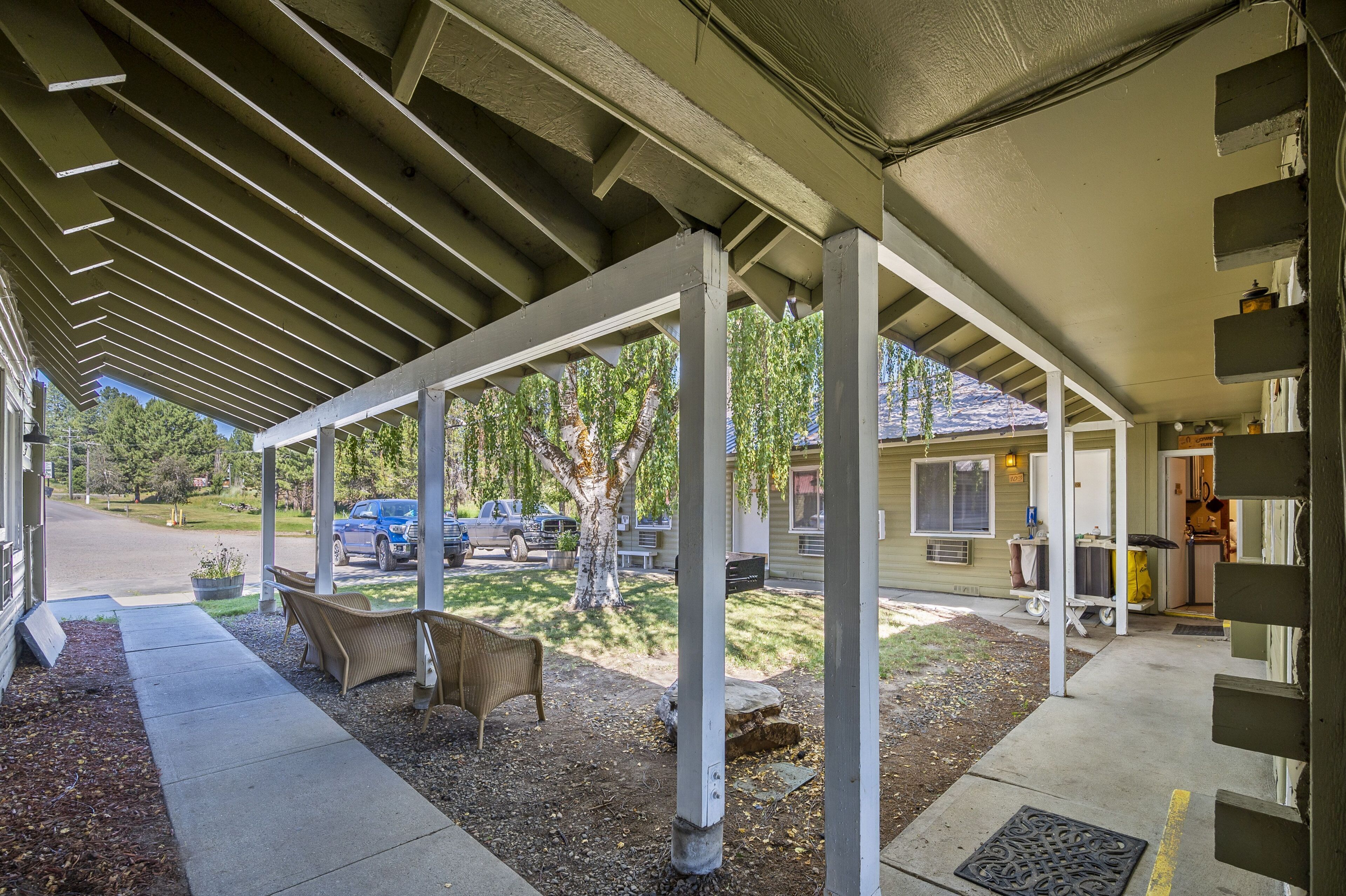 Un espace extérieur abrité, doté d’une structure en forme de pergola, d’un coin salon avec des chaises, et offrant une vue sur les véhicules garés et une maison en arrière-plan.