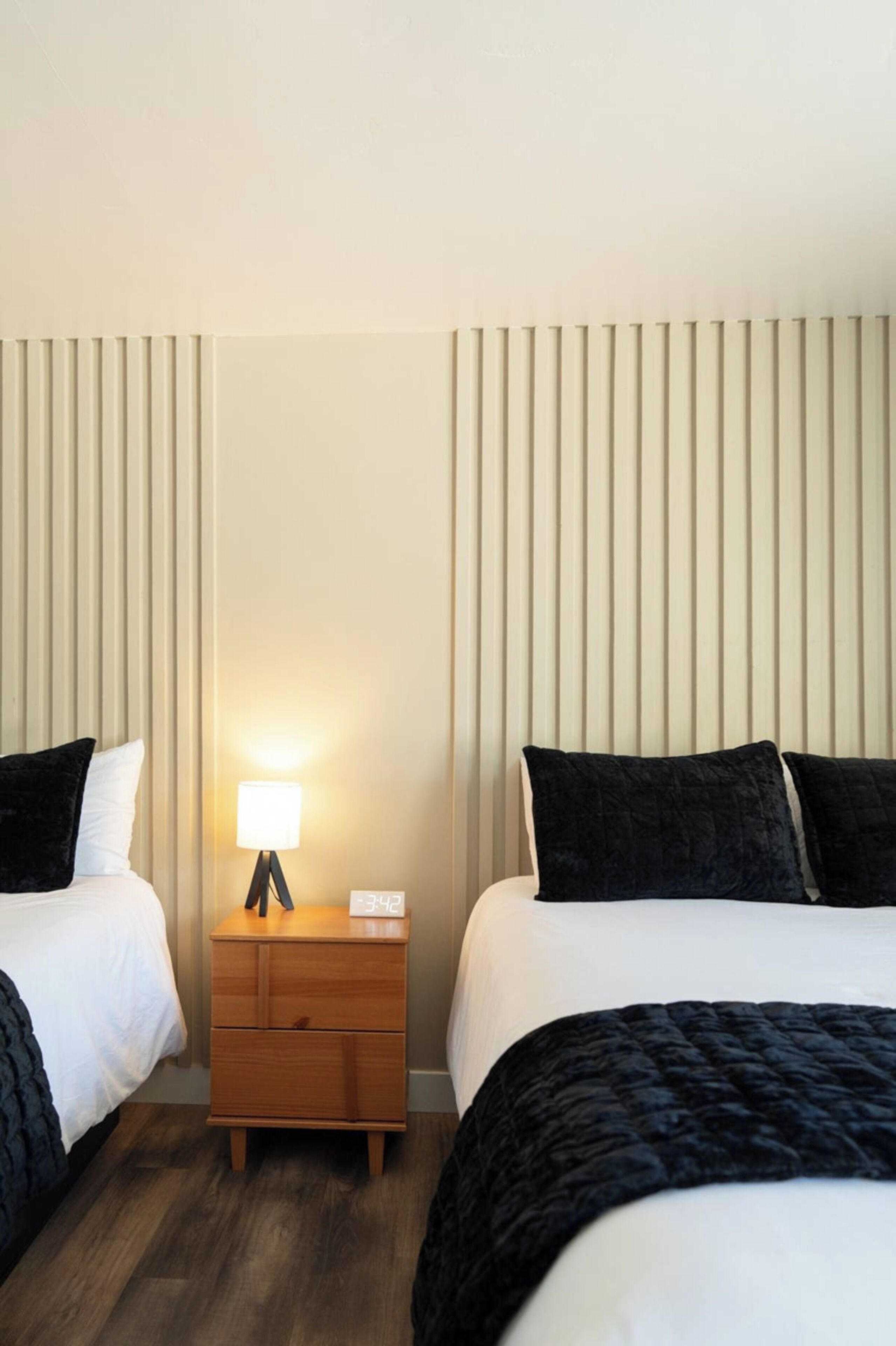 Ein Hotelzimmer mit zwei Betten, einem Nachttisch mit Lampe und senkrechten Jalousien an den Fenstern.