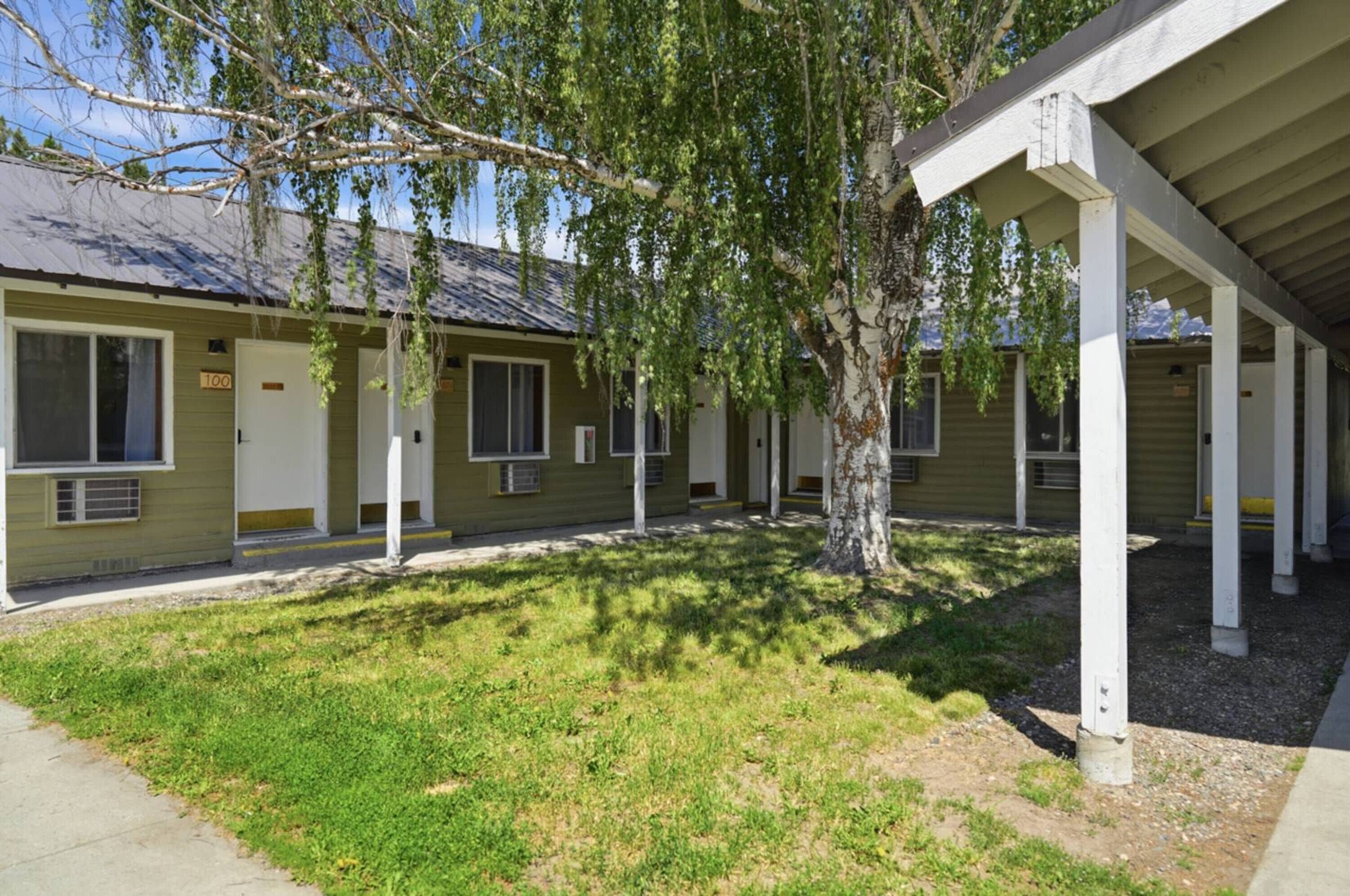 A row of motel-style buildings with individual rooms, a covered outdoor area, and a tree in the foreground.