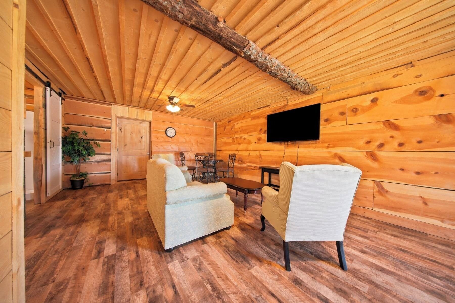 Interior de una cabaña de troncos con comedor, dos sofás, televisión y un reloj en la pared.