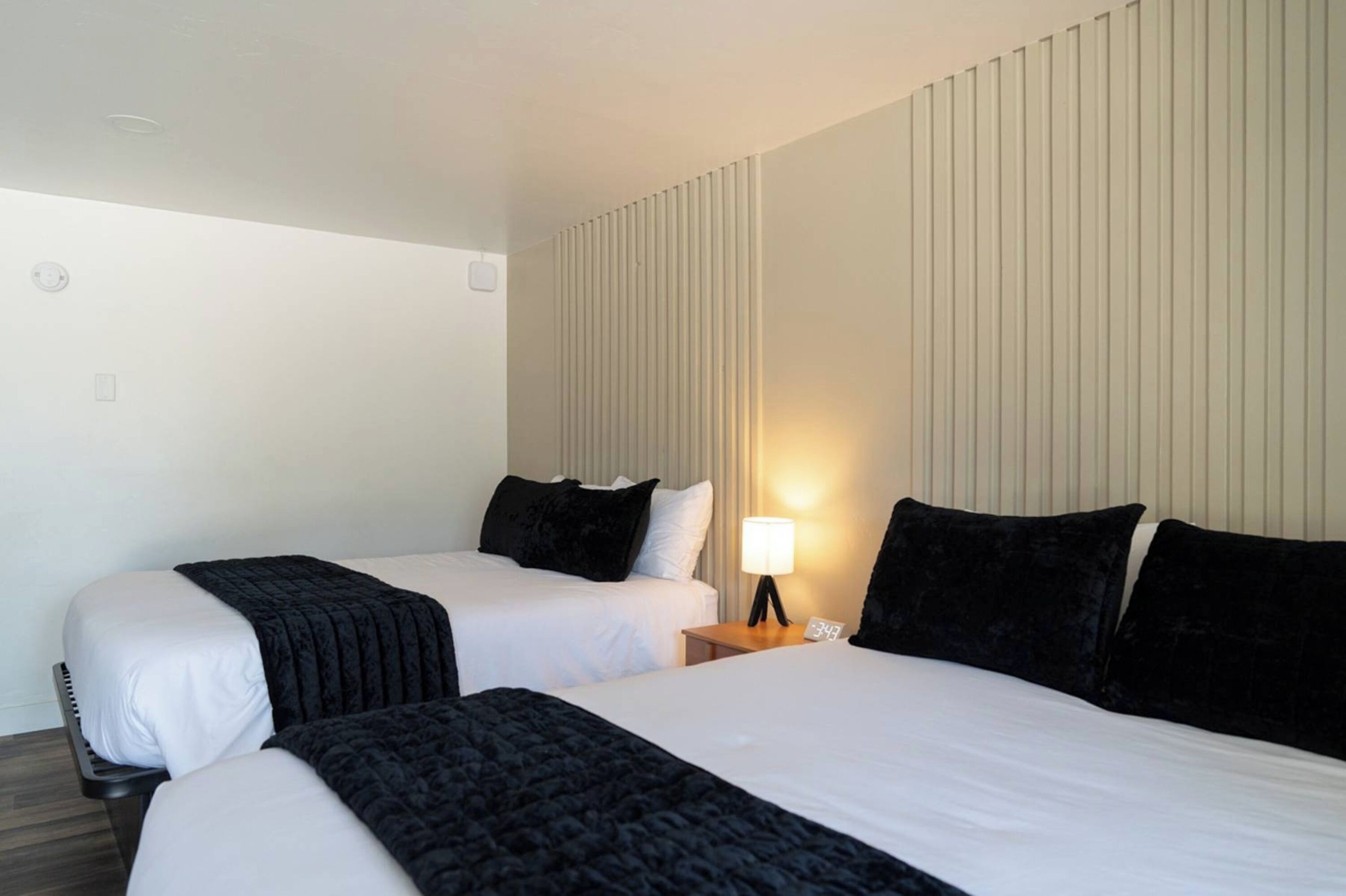 A hotel room with two beds, a nightstand with a lamp, and a wall-mounted control panel.